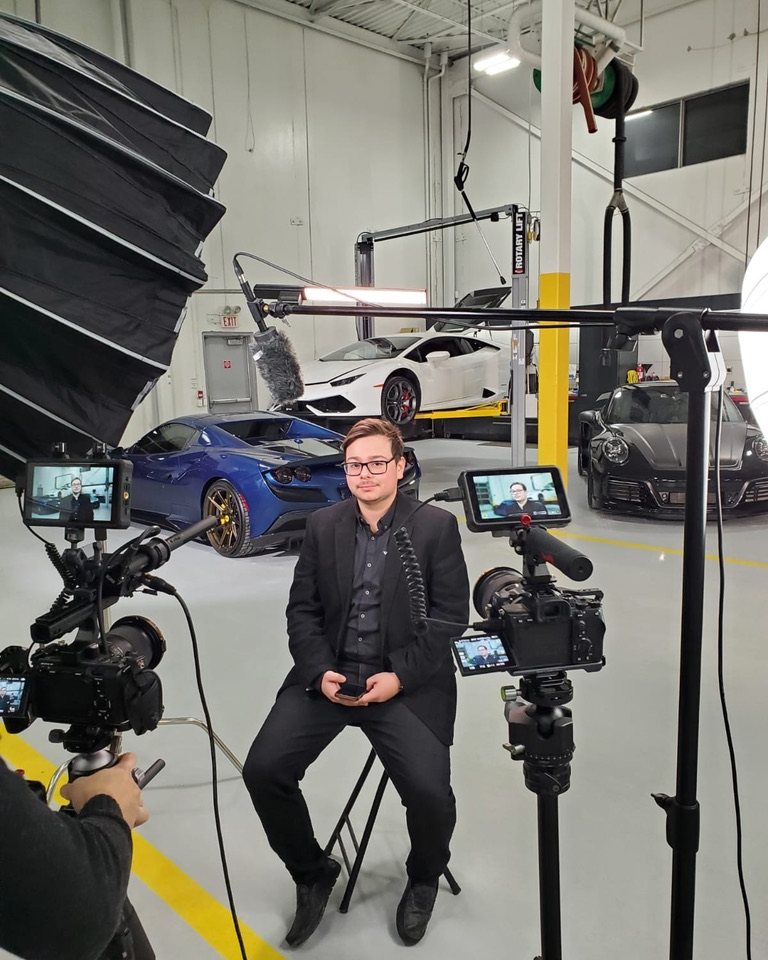 Juan in a black suit on a studio stool between two cameras, with supercars behind him